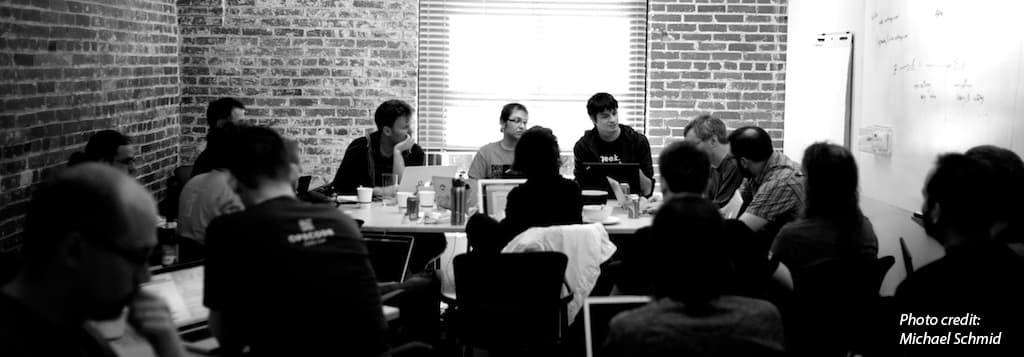 Numerous contributors sit around a table while others listen from behind.